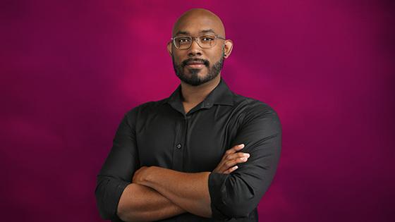 Gregg is wearing a black shirt and rectangular glasses. He is standing in front of a pink background with his arms crossed.